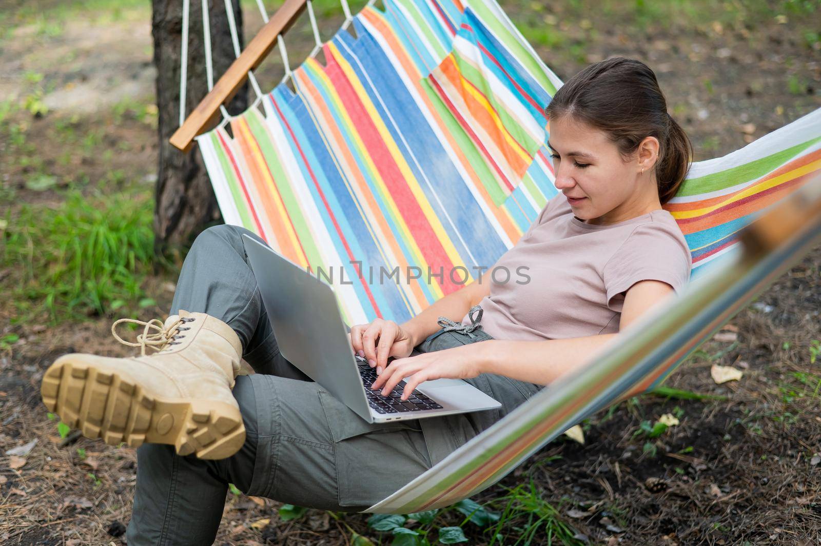 Caucasian woman working on laptop while sitting in a hammock in the forest. Girl uses a wireless computer on a hike