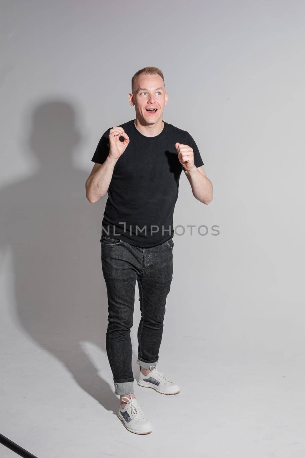 Full length of adult Caucasian blonde man in black t-shirt, dark jeans and sneakers talking to camera with arms bent, holding fists. Presenter or advertiser promoting something.