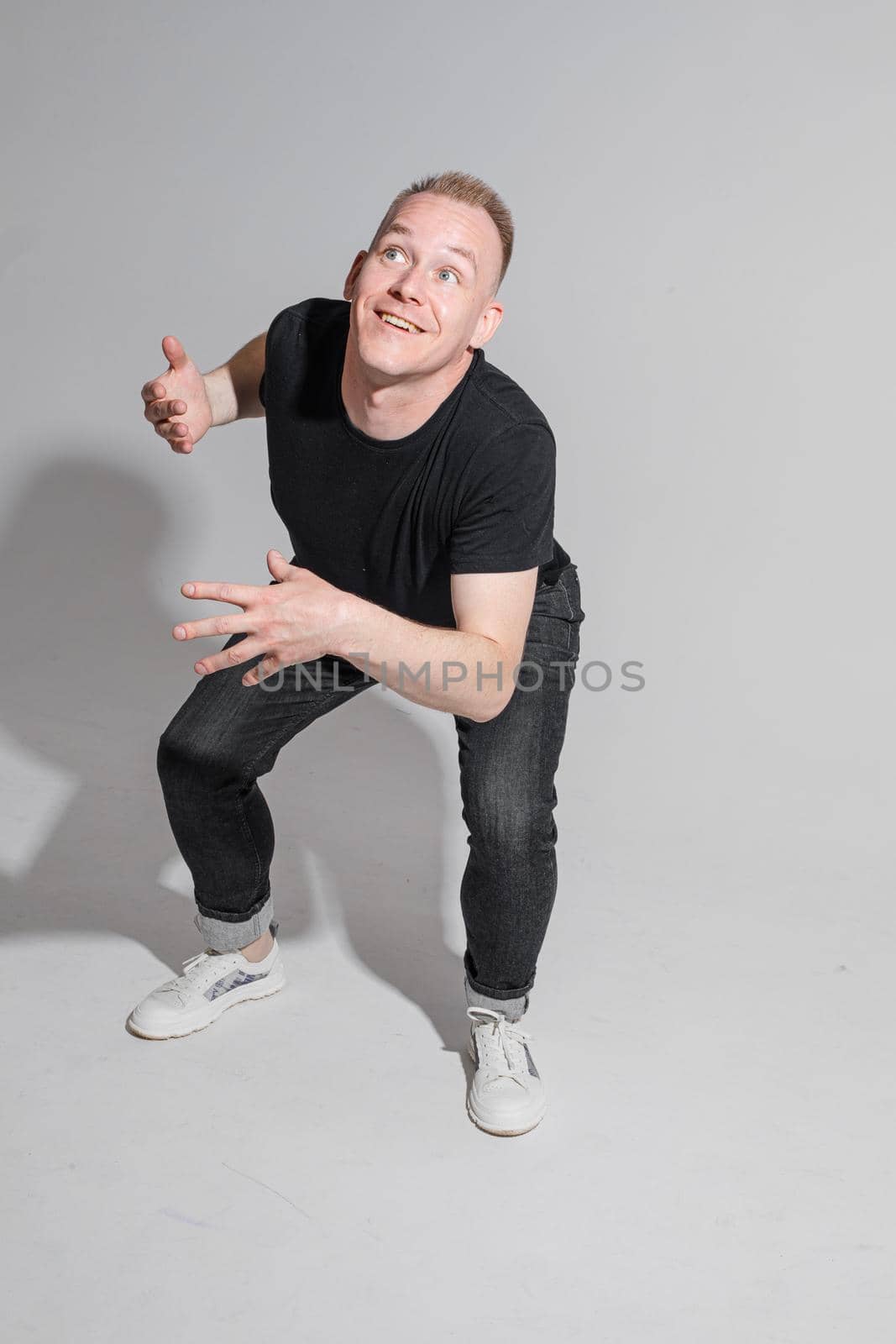 Full length of adult Caucasian blonde man in black t-shirt, dark jeans and sneakers talking to camera with arms bent, holding fists. Presenter or advertiser promoting something.