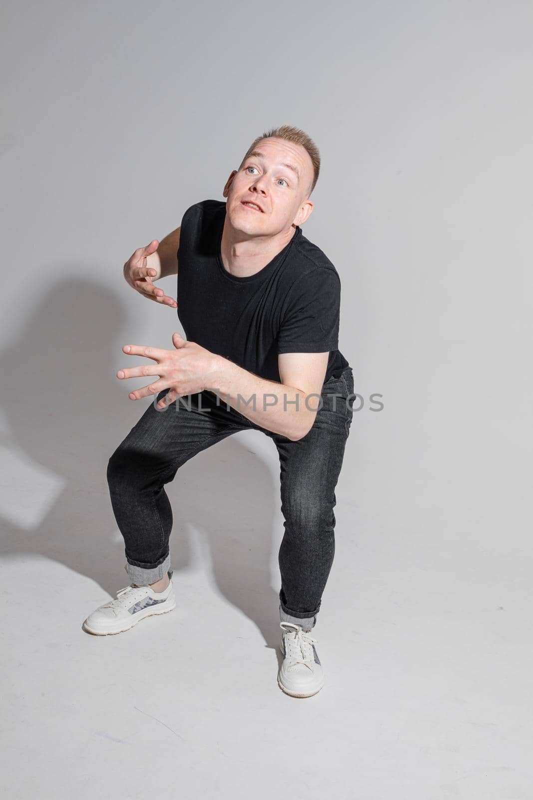 Full length of adult Caucasian blonde man in black t-shirt, dark jeans and sneakers talking to camera with arms bent, holding fists. Presenter or advertiser promoting something.