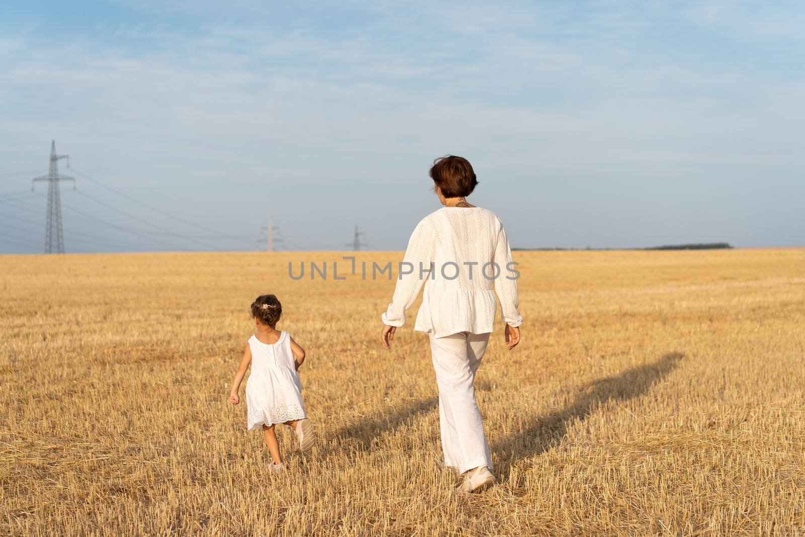 Little girl with her mother dressed white clothes walking wheat field back view blue sky background. Daughter with mom run nature countryside view Freedom or happy family concept