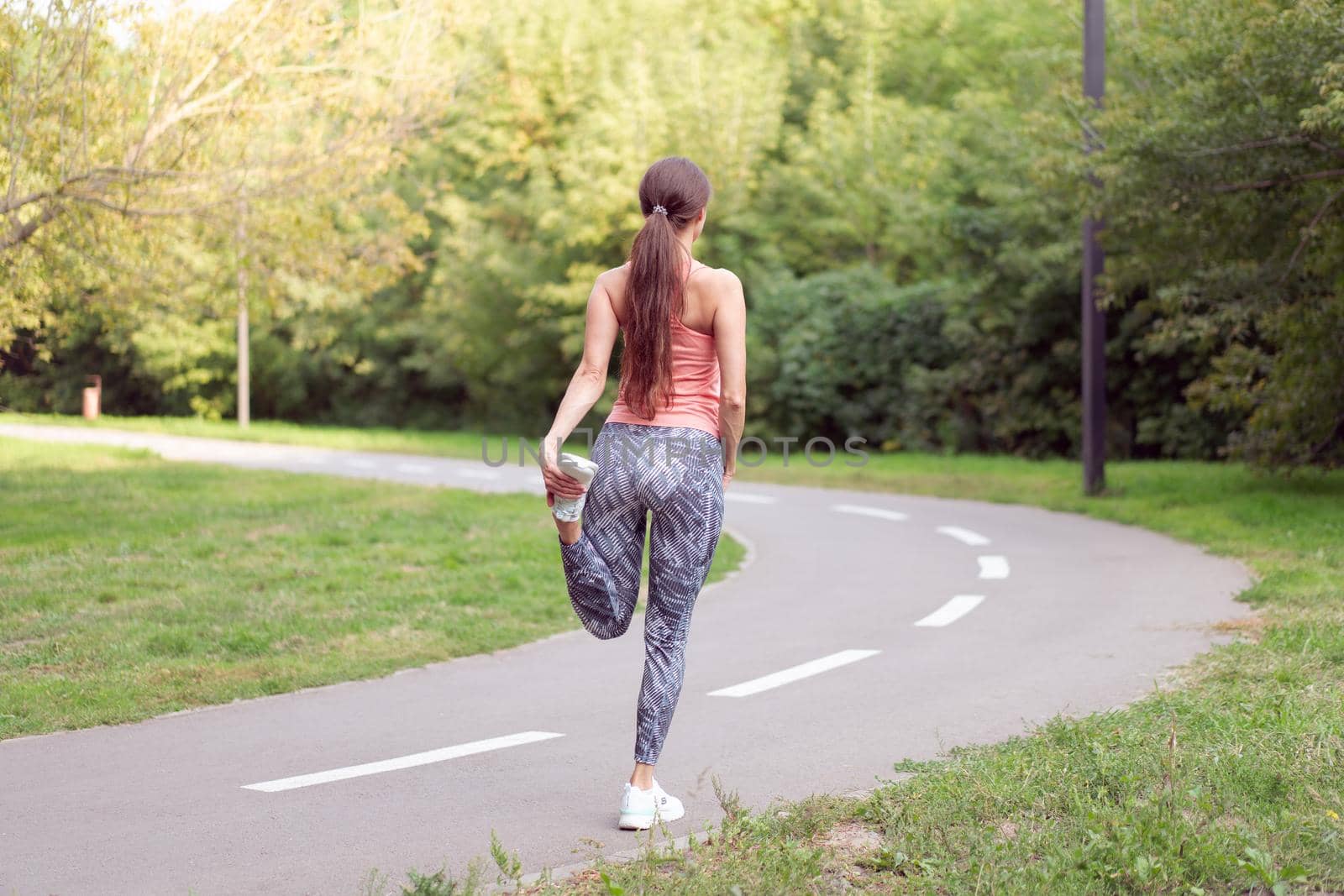 Athletic woman warming up legs before running exercises standing running track in summer park Morning cardio training Middle age caucasian female warm up body before jogging asphalt path in public park