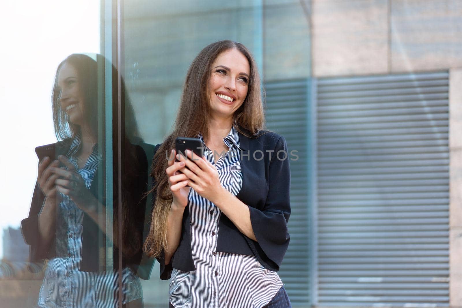 Business woman 35 years dressed stripe shirt black jacket with long hair standing near office building outdoor hold smartphone in hand Happy Caucasian female business person use technology