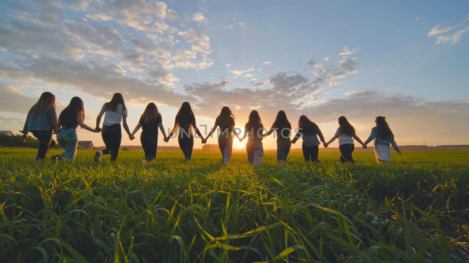 A group of girls walk towards the sun at sunset holding hands