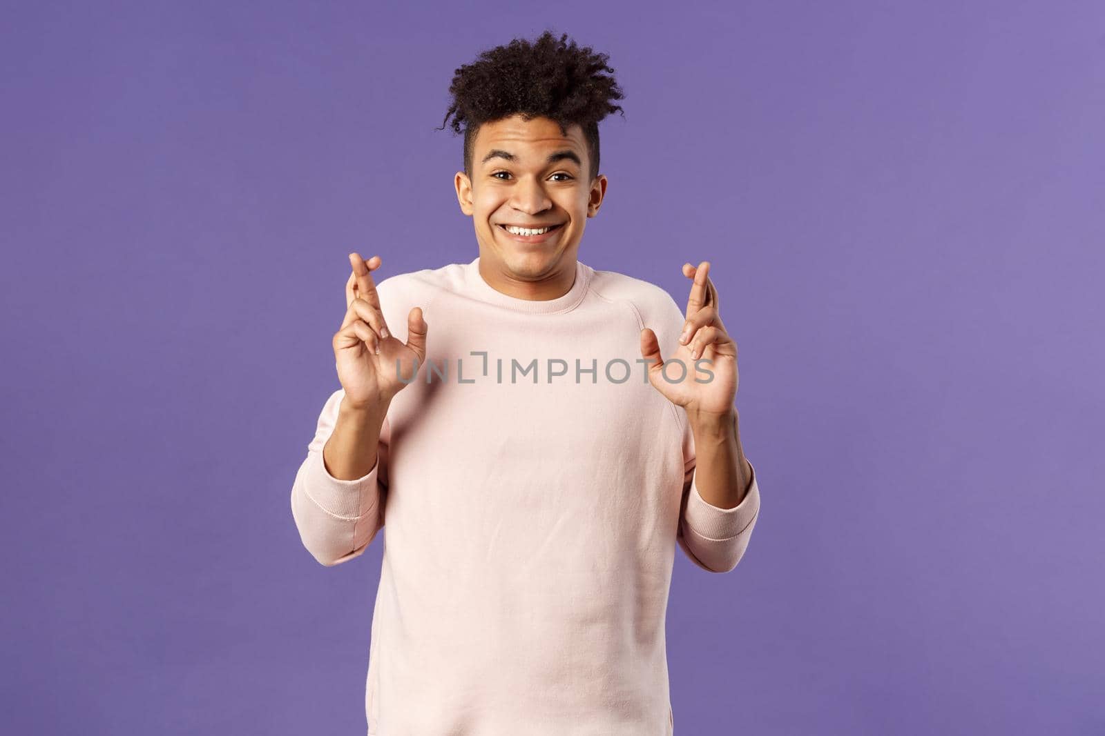 Portrait of hopeful positive, smiling young man having faith in dreams may come true, make wish, cross fingers good luck and awaiting for miracle, standing purple background praying.