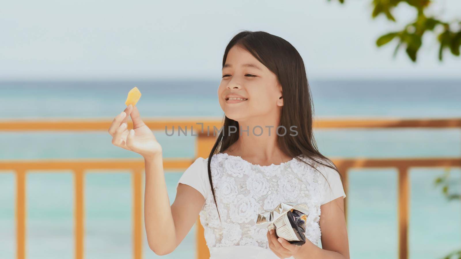 Philippine schoolgirl in white dress smiles, eating crispy potato chips. Tropical landscape. The palms. Summer