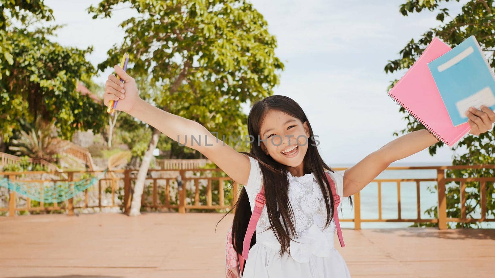 A charming philippine schoolgirl with a backpack and books in a park off the coast. A girl joyfully poses, raising her hands up with textbooks in her hands. Warm sunny day. by DovidPro