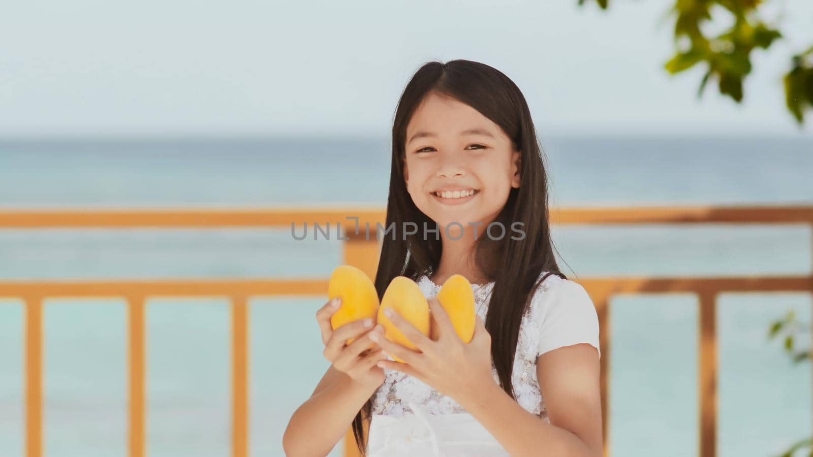 A charming philippine schoolgirl girl in a white dress and long hair positively poses with a mango in her hands. The sun. The blue ocean. Childhood. Recreation. by DovidPro