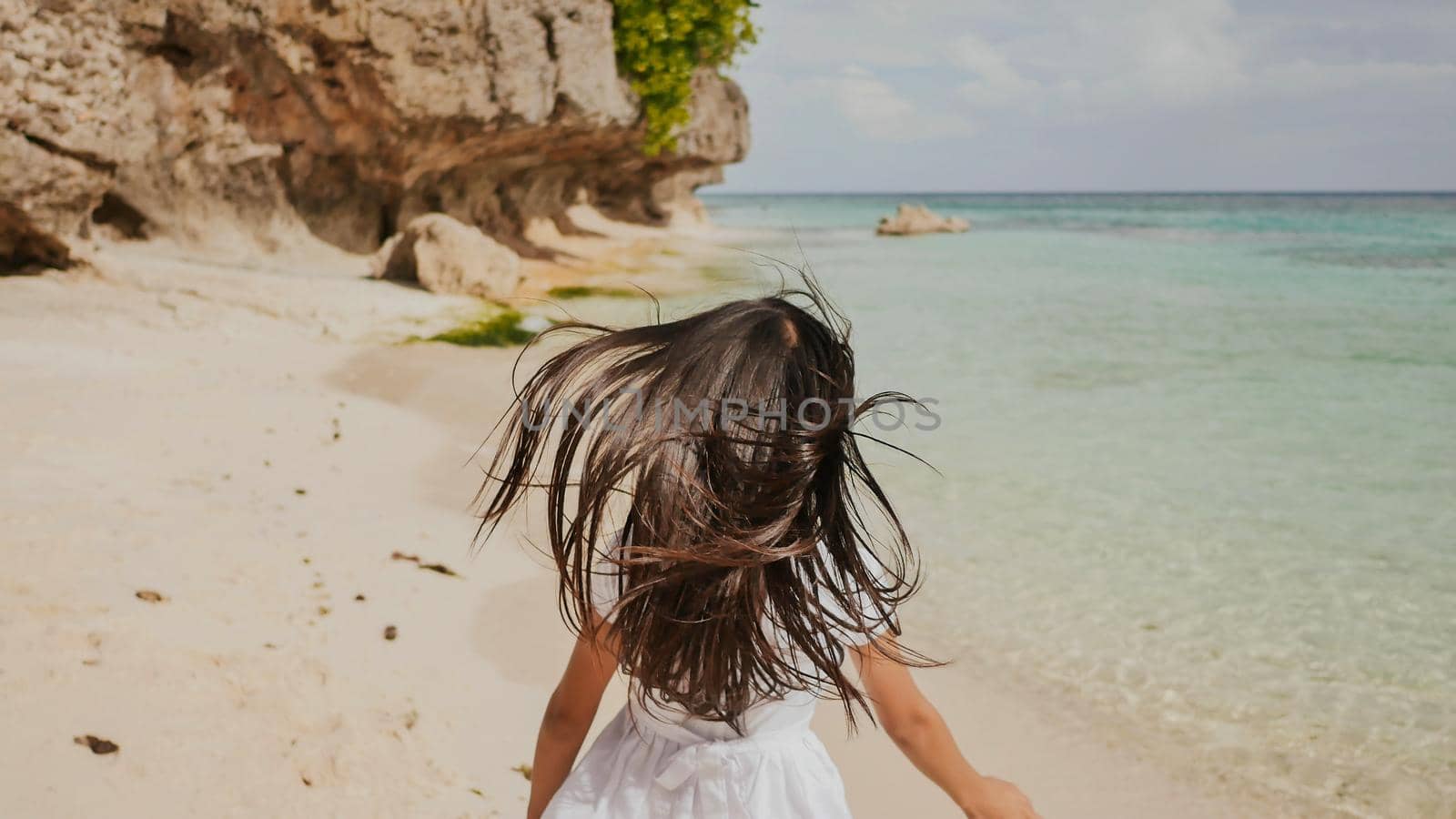 A charming and happy philippine teenage girl in a white summer dress is running along a tropical beach near the rocks. She is happily spinning. Childhood. Recreation