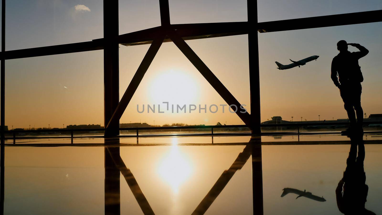 Silhouette of a tourist guy watching the take-off of the plane standing at the airport window at sunset in the evening. Travel concept, people in the airport