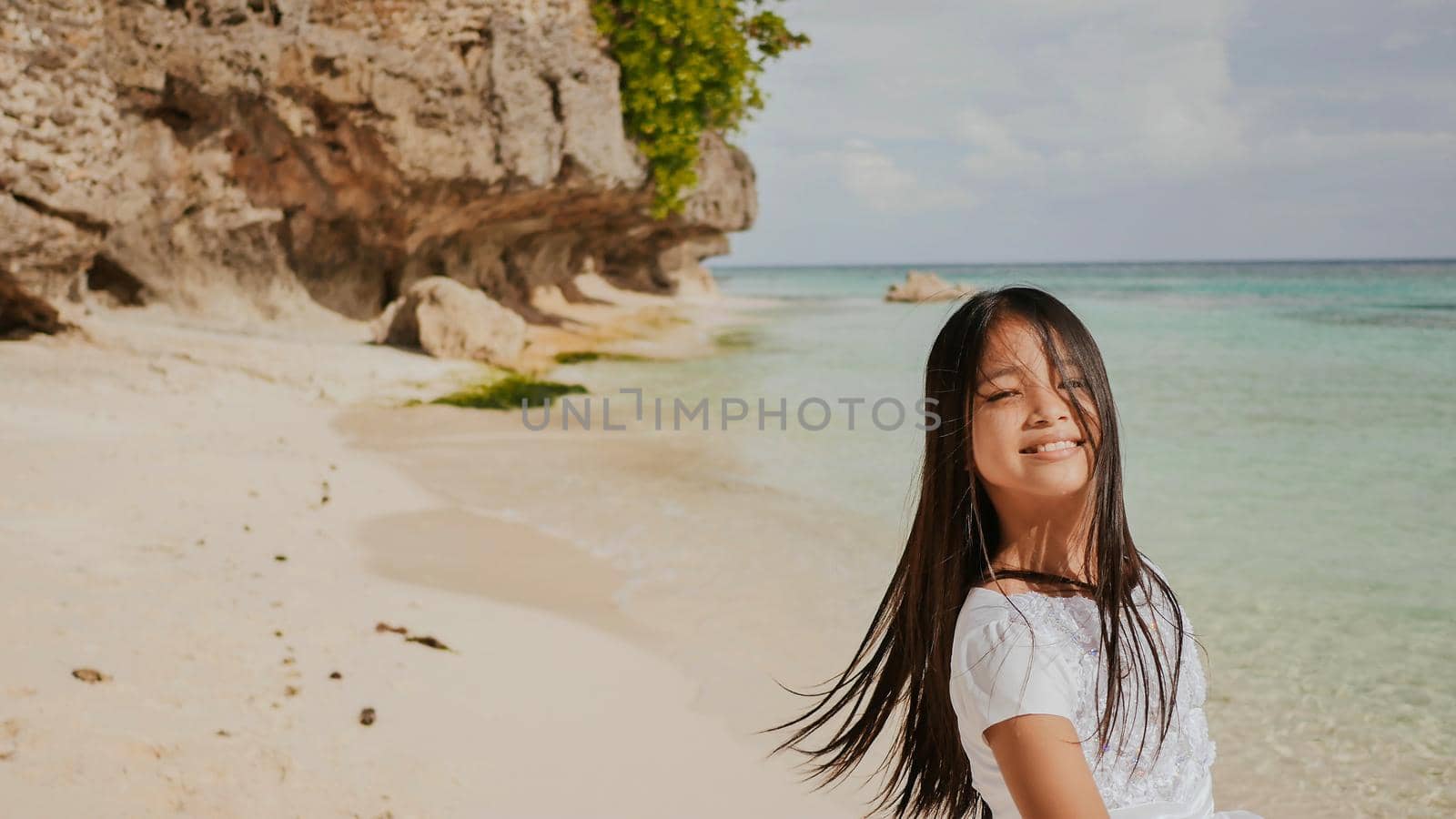 A charming and happy philippine teenage girl in a white summer dress is running along a tropical beach near the rocks. She is happily spinning. Childhood. Recreation