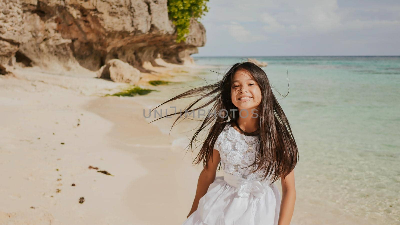 A charming and happy philippine teenage girl in a white summer dress is running along a tropical beach near the rocks. She is happily spinning. Childhood. Recreation