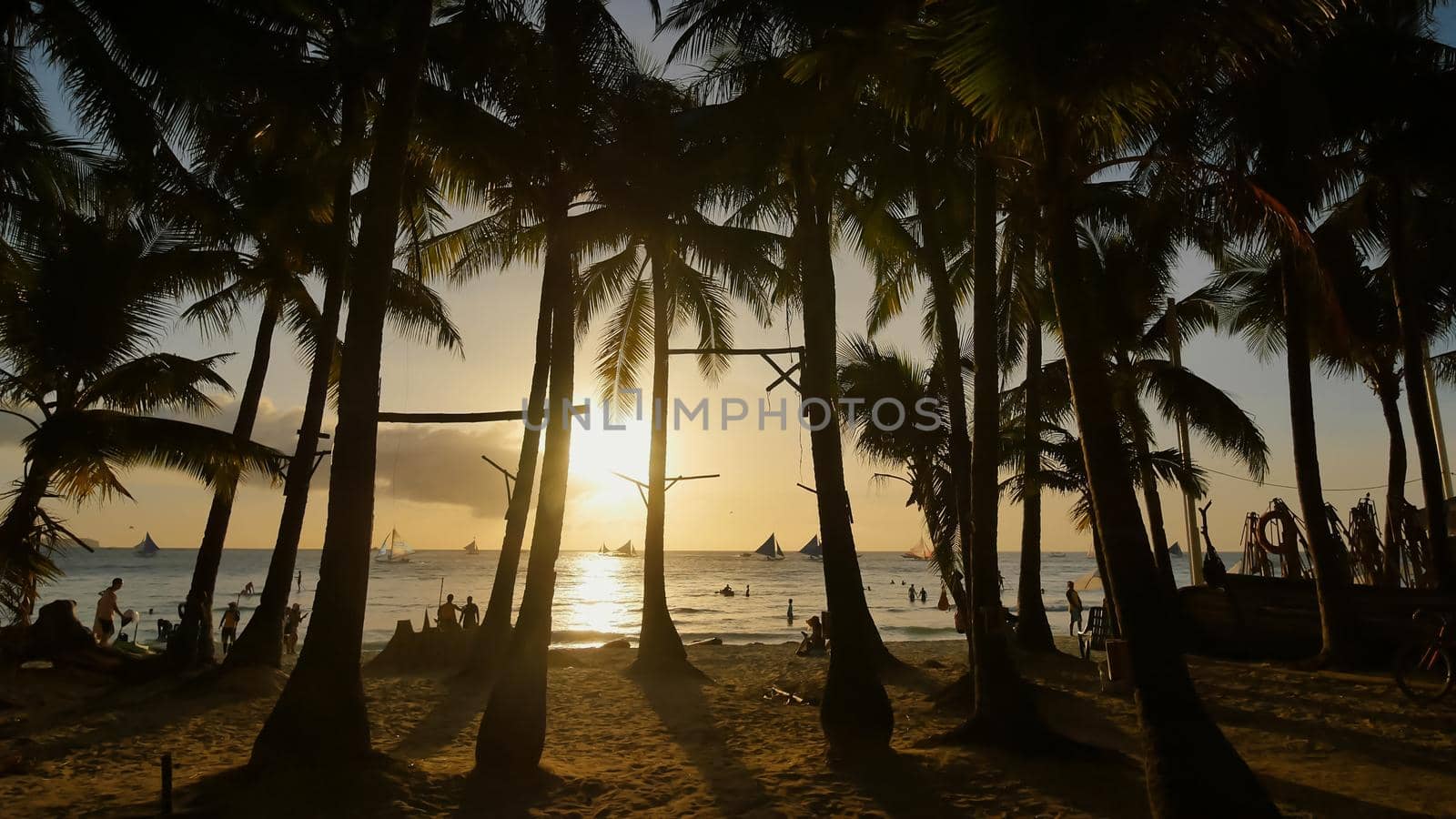 Beach with silhouettes of tourists among palm trees on the island of Boracay. Palm trees in the rays of sunset. Sailboats on the water. Philippine Tropics