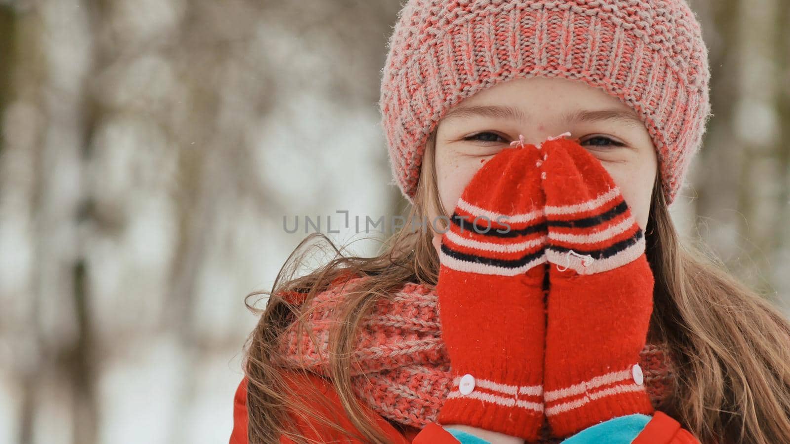 Portrait of a young schoolgirl with freckles in the woods in winter. He warms his hands in mittens and applies them to his face and lips. Shows movement hand in hand. by DovidPro