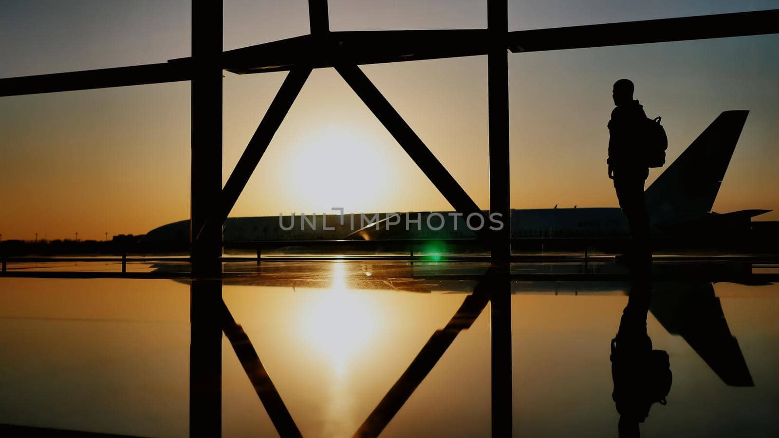 The silhouette of the tourist guy watching the takeoff of the plane and passing a huge plane standing at the airport window at sunset in the evening. The travel concept, people in the airport