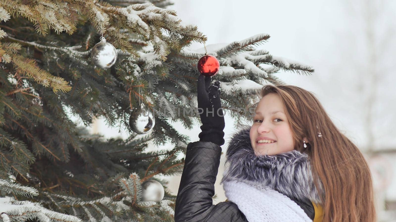 Attractive young girl with long hair in a winter suit posing against a snowy tree. A girl is stringing Christmas balls on a branch. Winter in the forest