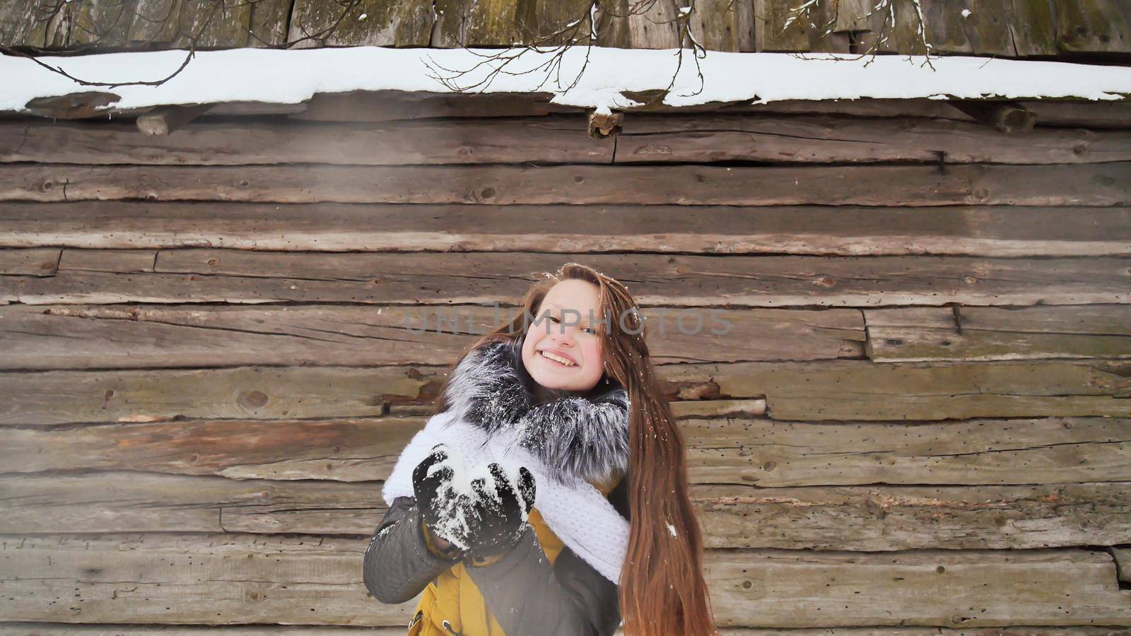 Young beautiful girl having fun blowing snow on the camera in winter clothes on the background of a wooden house in the village. Winter fun