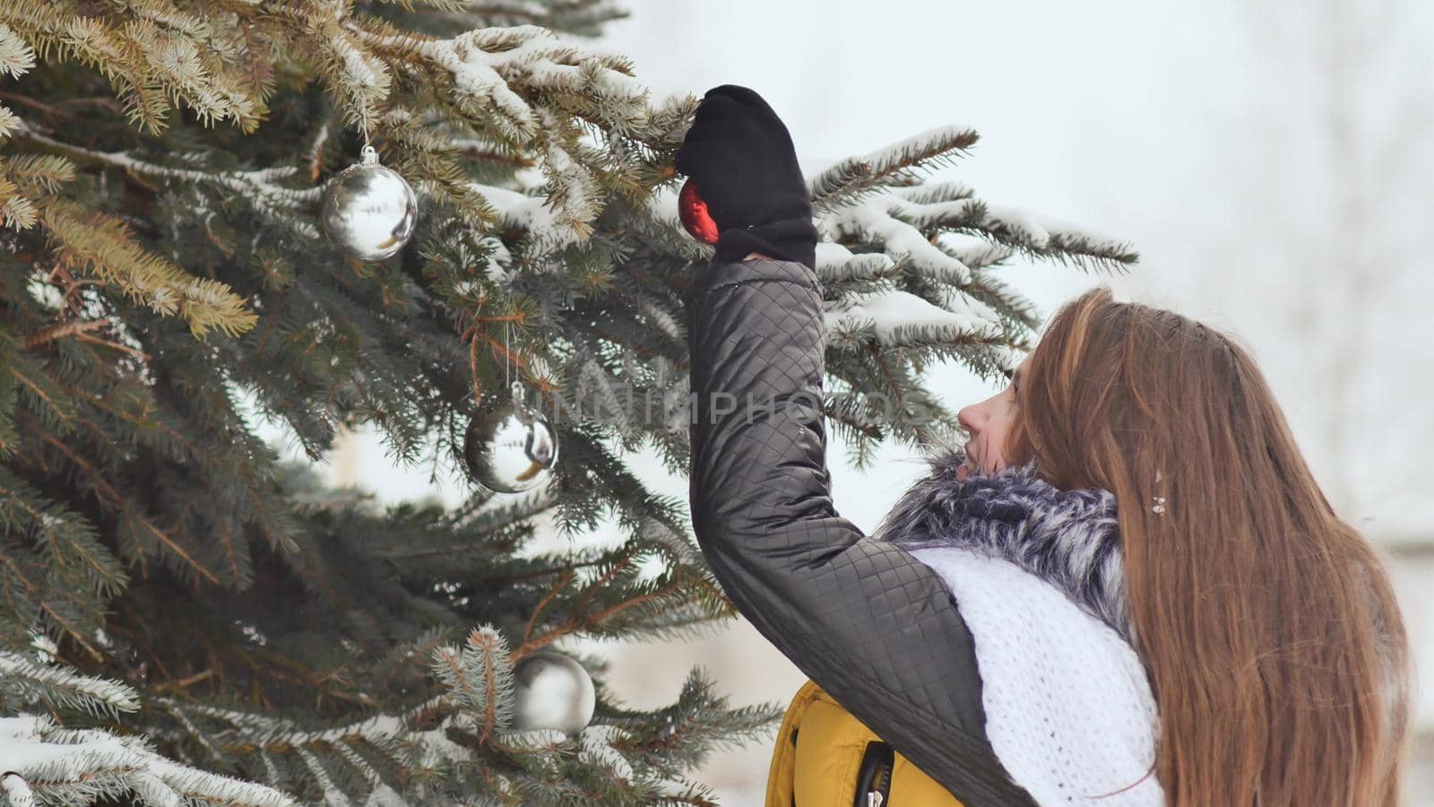 Attractive young girl with long hair in a winter suit posing against a snowy tree. A girl is stringing Christmas balls on a branch. Winter in the forest