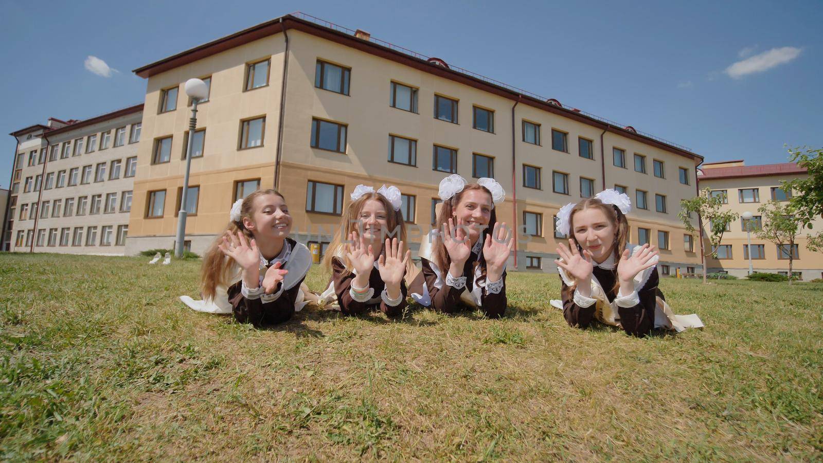 Four schoolgirl graduates are lying on the grass against the backdrop of their school. Girls waving their hands and fleeing. Russian school