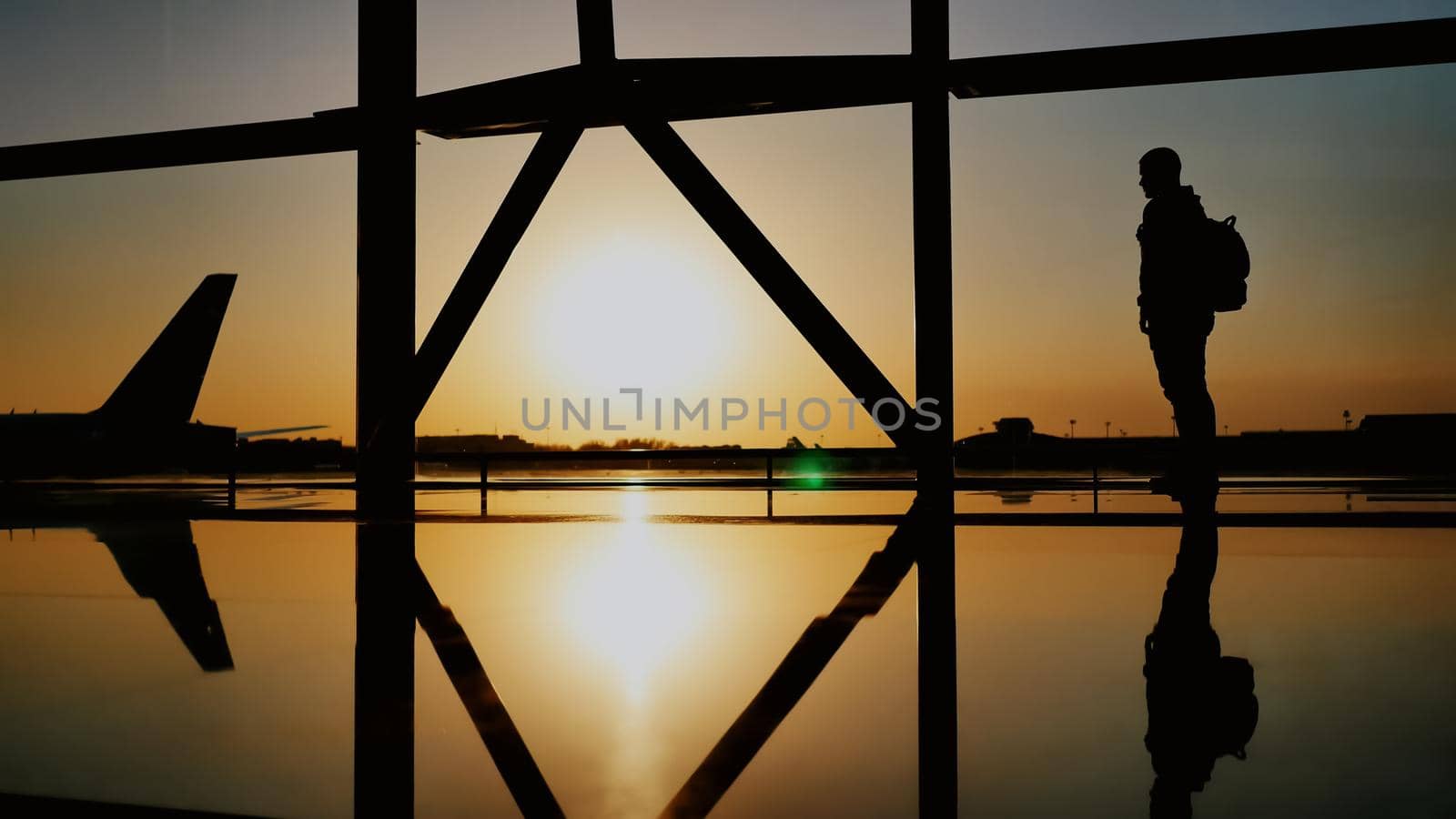 The silhouette of the tourist guy watching the takeoff of the plane and passing a huge plane standing at the airport window at sunset in the evening. The travel concept, people in the airport