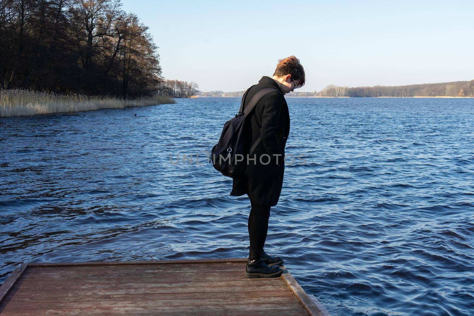 girl in a black coat stands at the edge of the pier and looks into the water of the lake