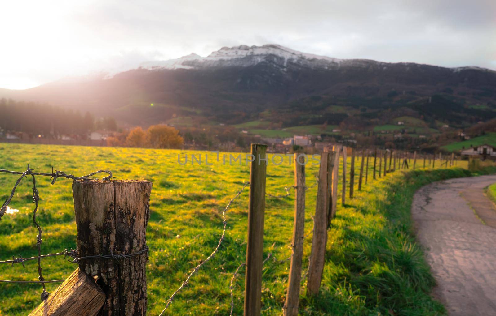 Old wooden pole of wire fence on blur green grass field, city in valley, and mountain in Europe. Fence of green animal grazing pasture beside the road. Wooden pole of green grass field with sunlight.