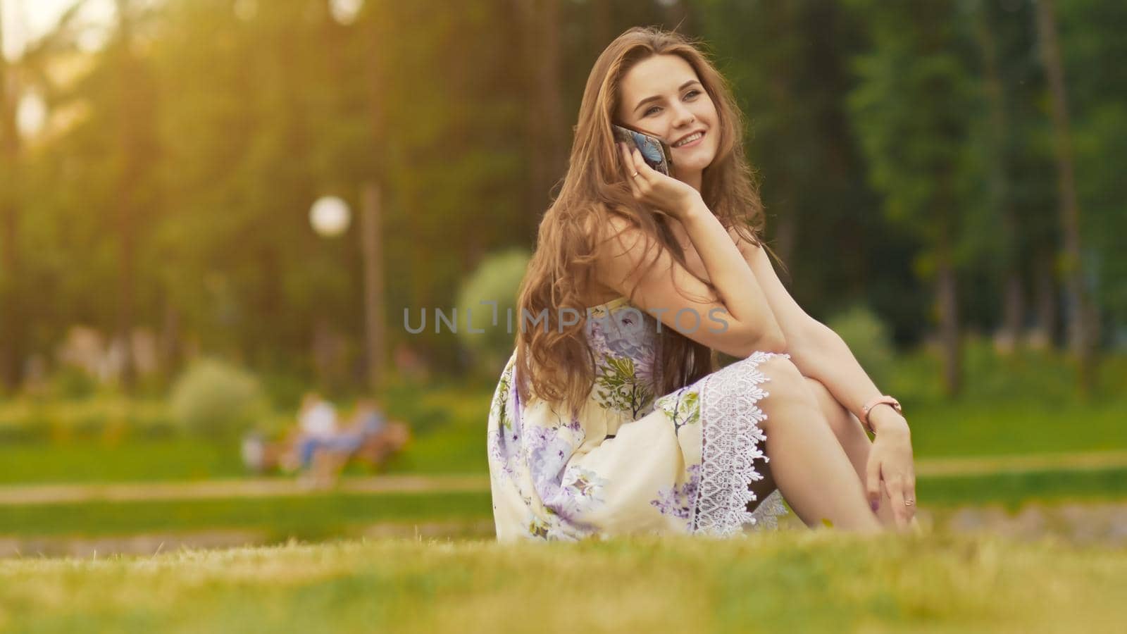 Young beautiful woman in summer dress with long hair sitting on grass in green park and talking on the phone, smiling. Summer. Recreation. Youth