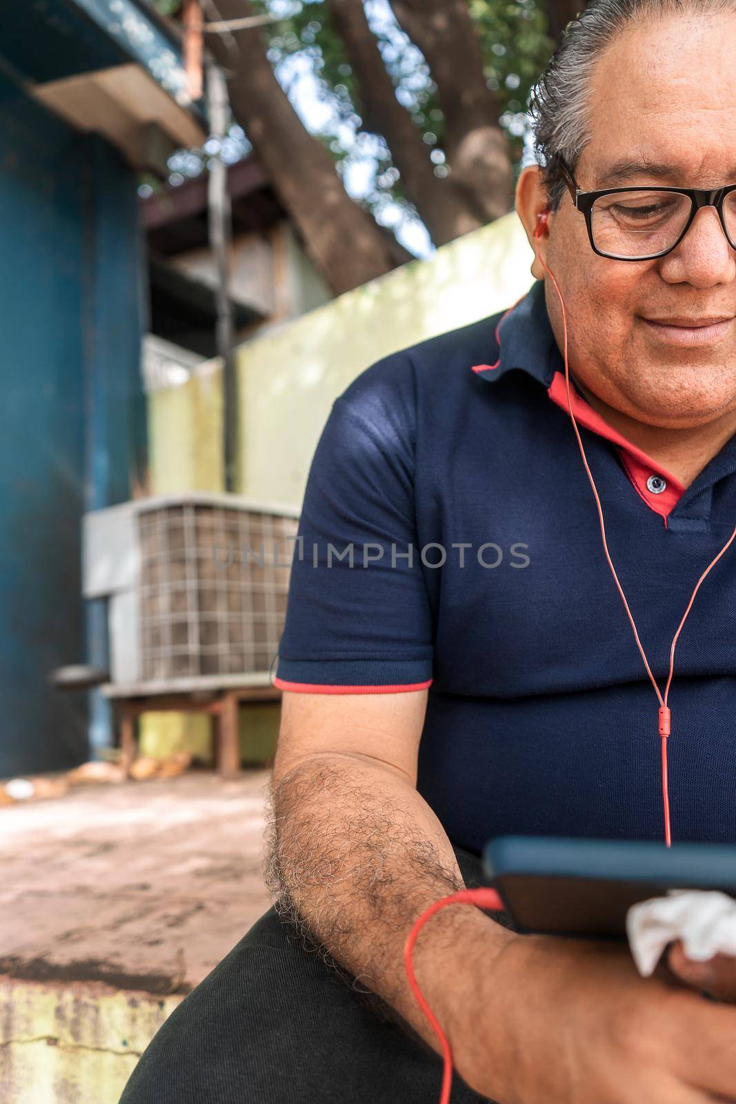 Vertical photo of a mature Latino man in glasses and shaved smiling looking at his cell phone outdoors
