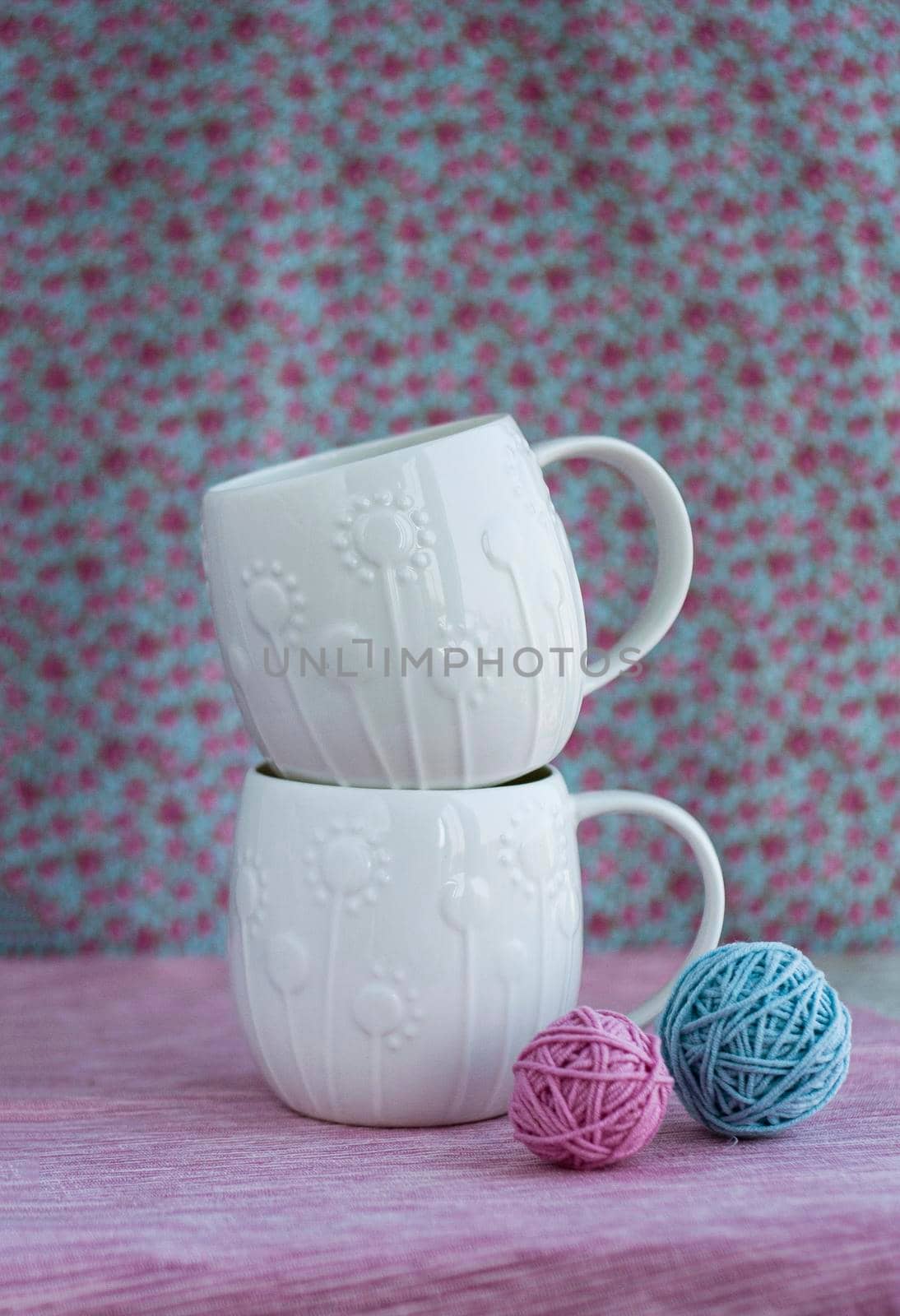 Two white cups stand on top of each other on a pink background, Nearby are two tangles for knitting pink and blue.
