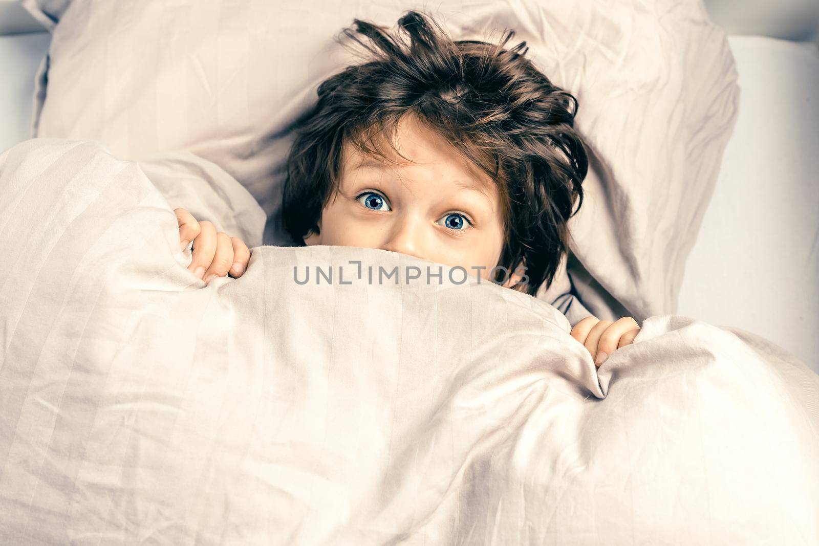 Surprised boy with funny face expression hiding and peeking from a duvet. Kid covering half of his face with blanket. Scared child, afraid of night monsters