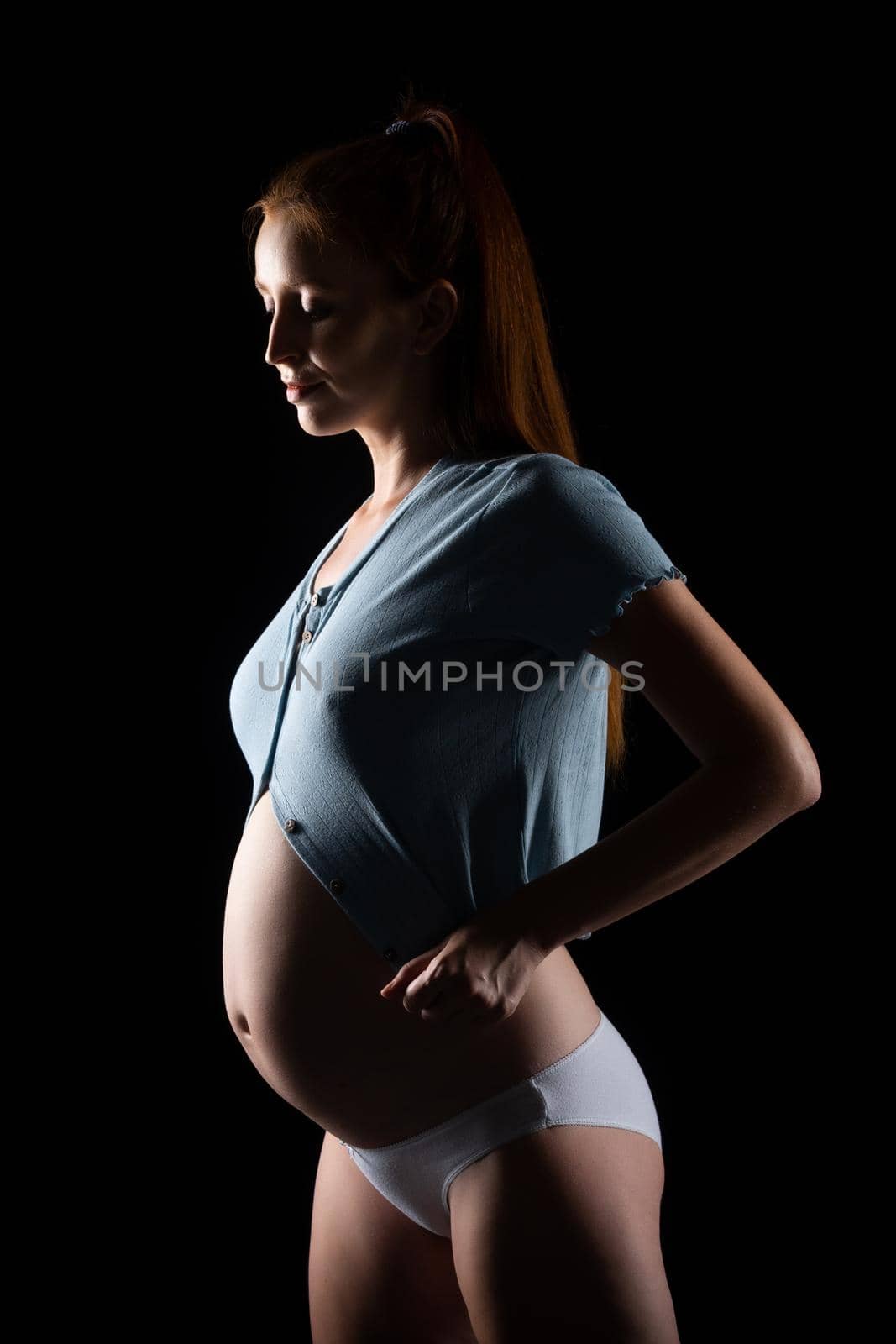 A pregnant woman with a big belly and red hair. Posing on a black background in the studio.