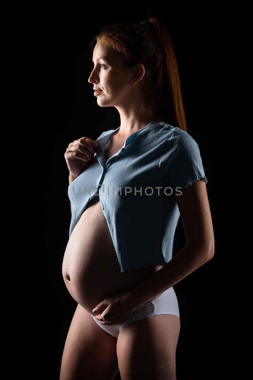 A pregnant woman with a big belly and red hair. Posing on a black background in the studio.