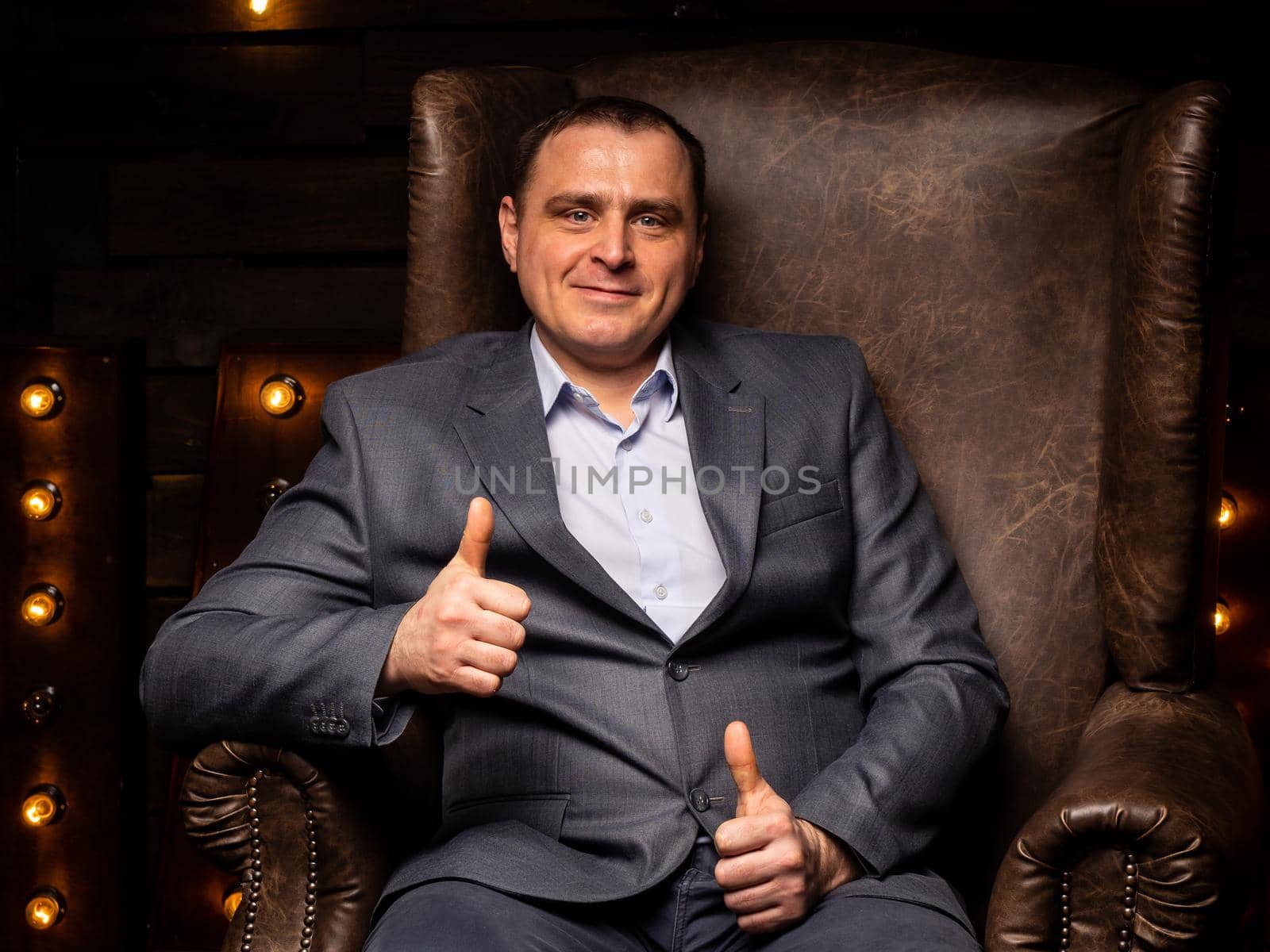 handsome businessman in a suit looks at the camera on a leather chair. by Evgenii_Leontev