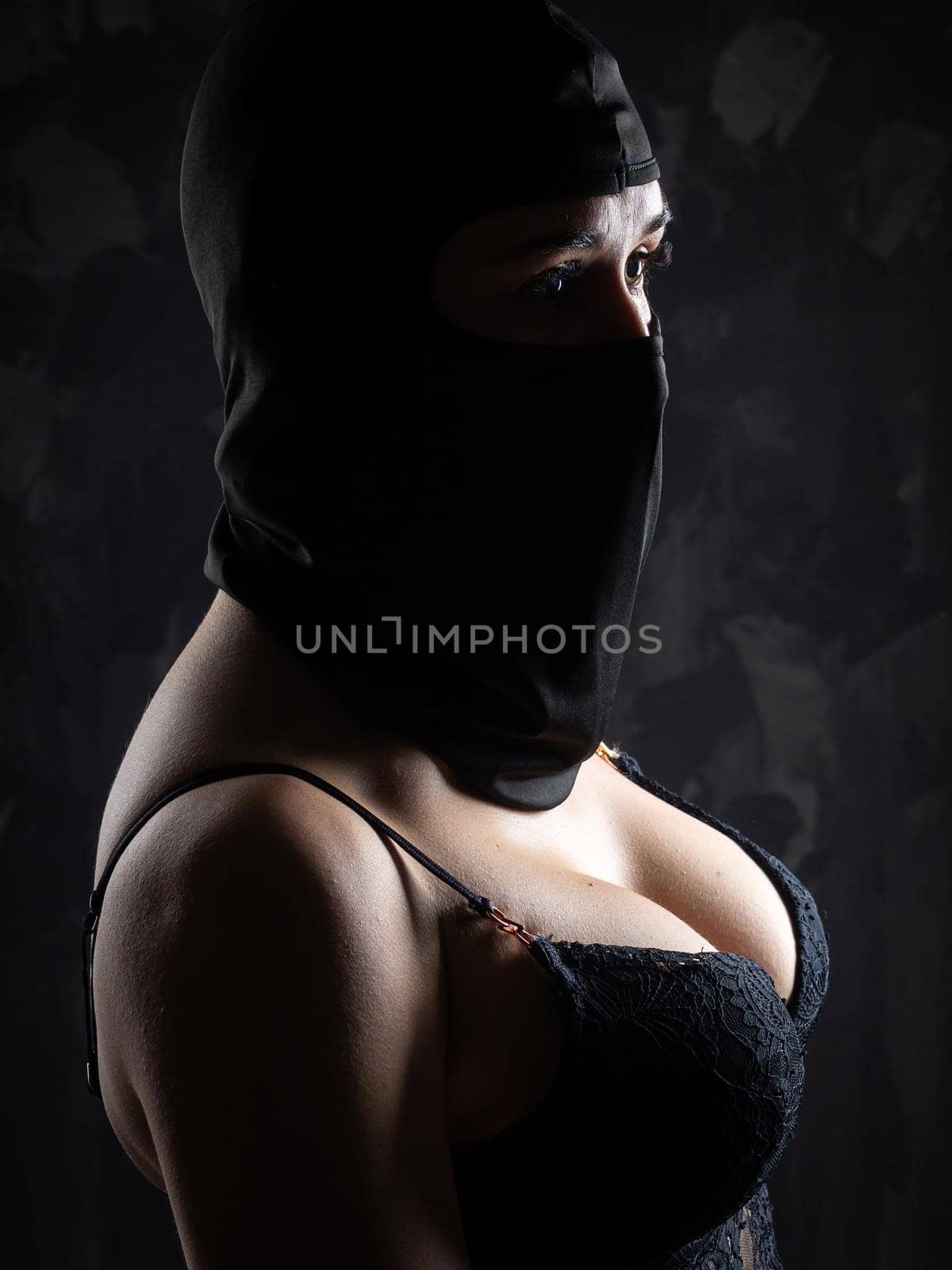 Portrait of a girl in a black balaclava and a black bra. Shot in the studio on a dark background.