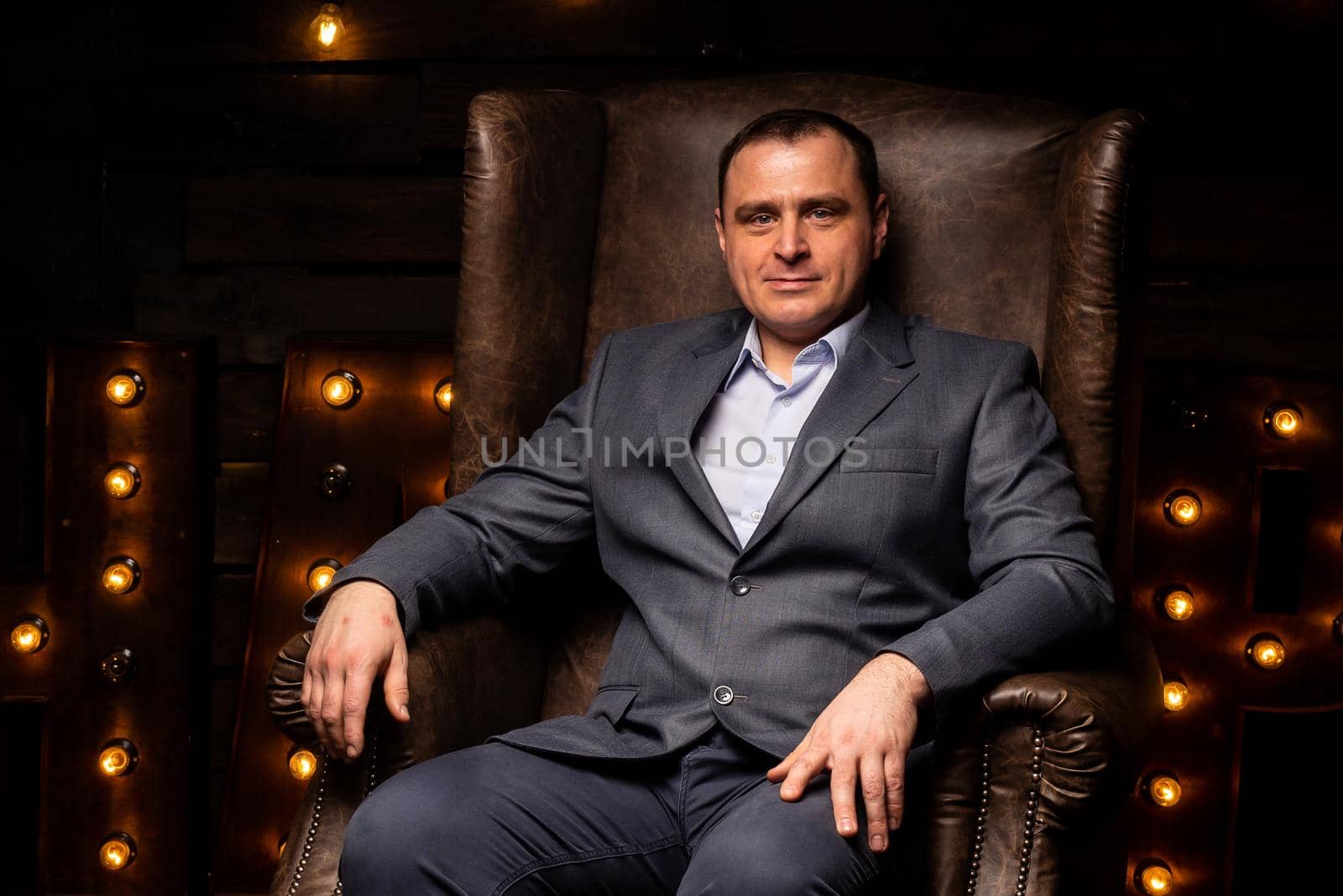 handsome businessman in a suit looks at the camera on a leather chair. by Evgenii_Leontev