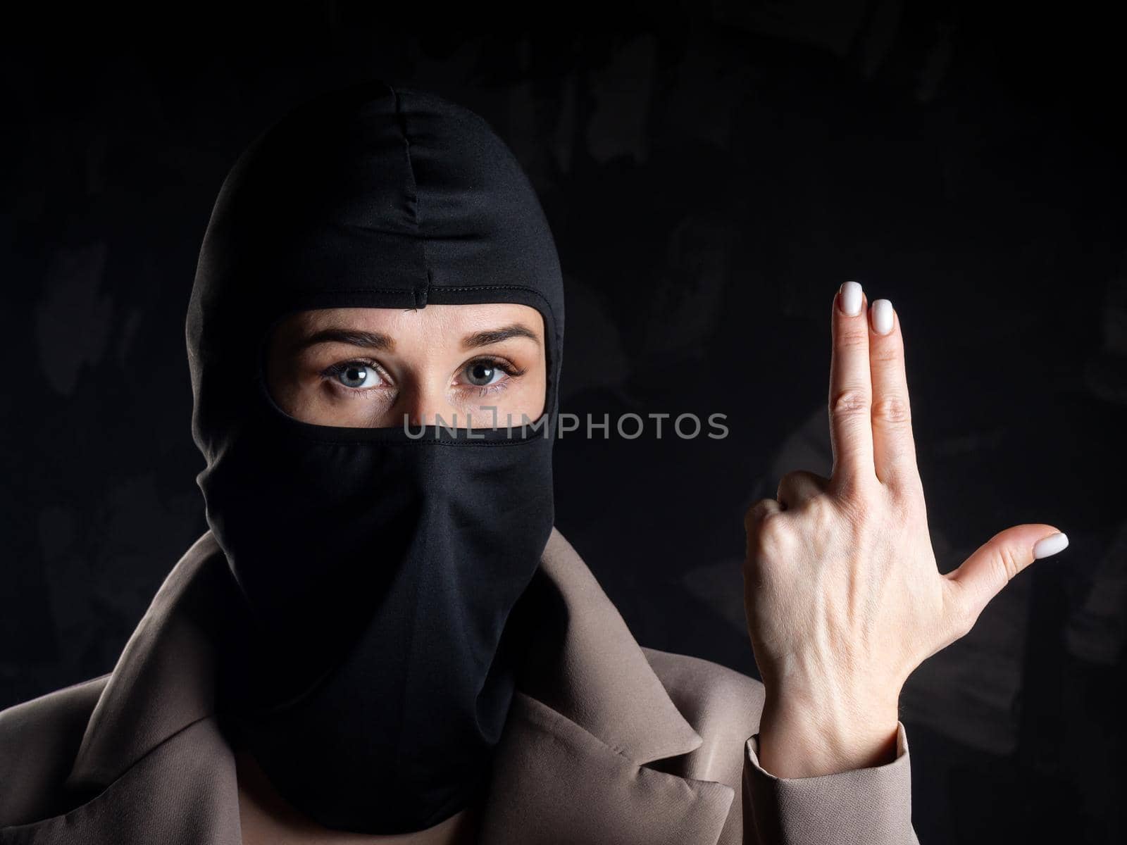 Portrait of a girl in a black balaclava and beige coat. Shot in the studio on a dark background.