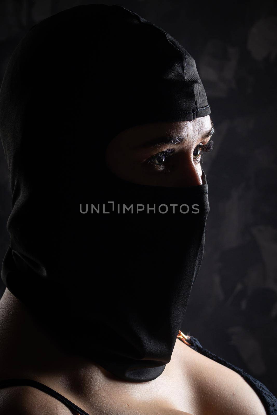 Portrait of a girl in a black balaclava and a black bra. Shot in the studio on a dark background.