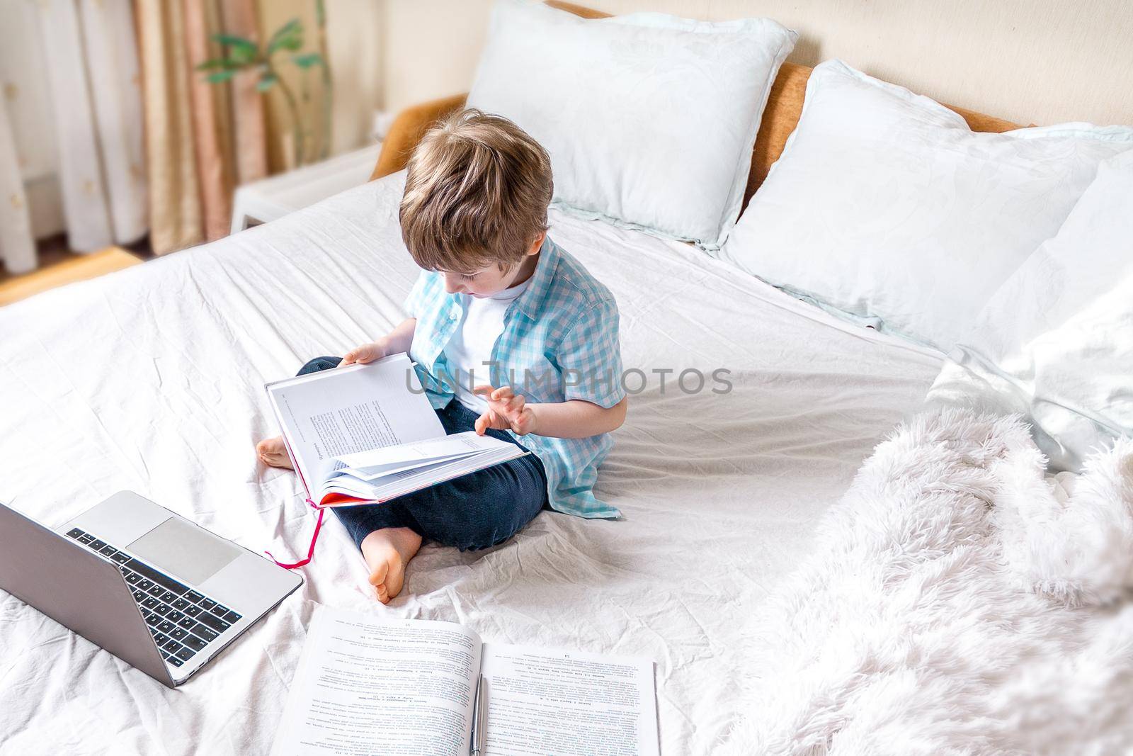 Distance learning online education. Caucasian boy with book studying at home and doing school homework. Sitting on bed with training books.