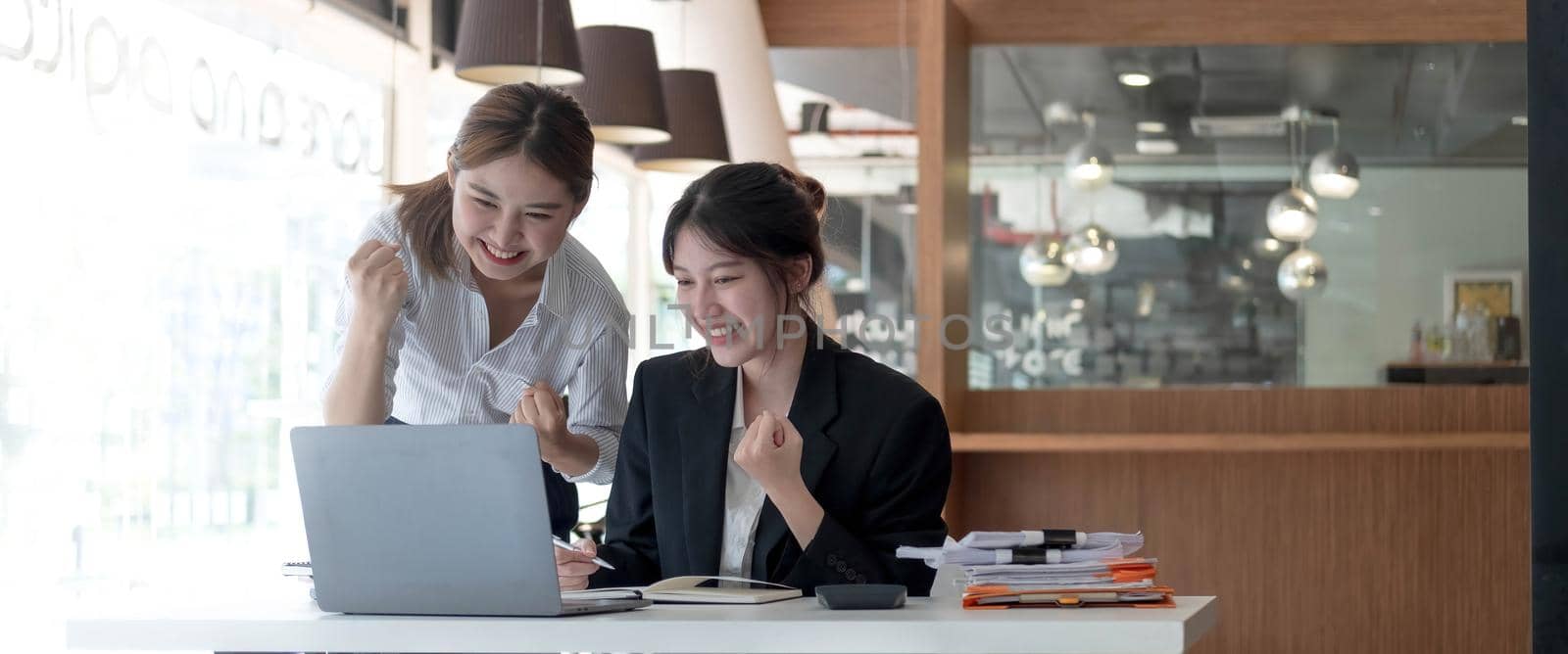 Two young Asian businesswomen show joyful expression of success at work smiling happily with a laptop computer in a modern office..