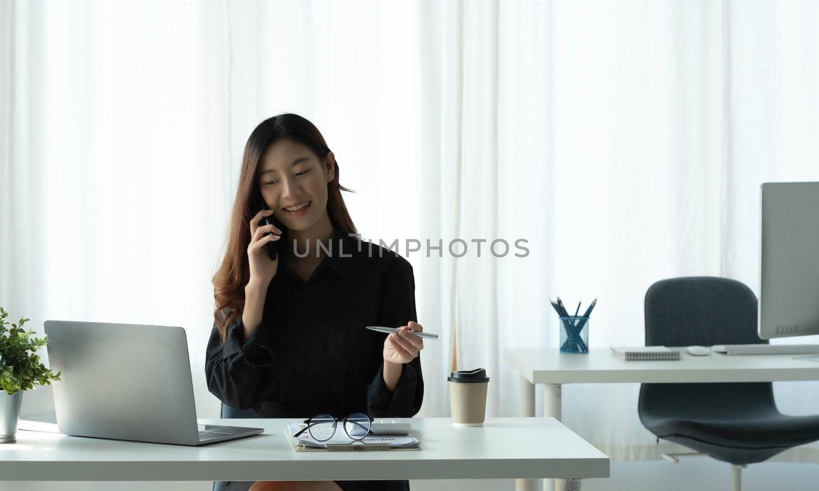 Smiling young Asian woman talking on the phone and take notes working at office.