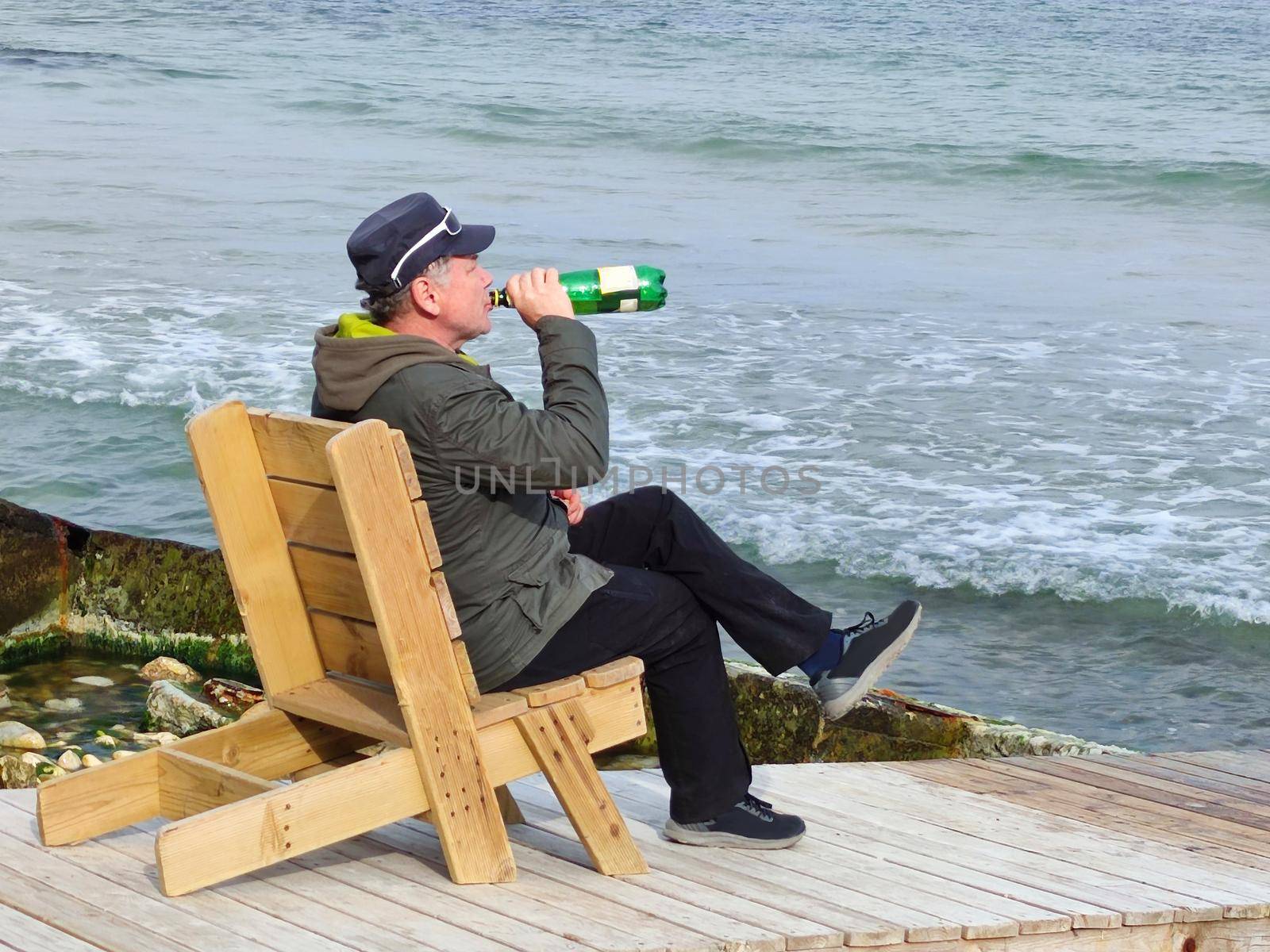 a man drinking beer sitting on a wooden deck chair by the sea by Annado