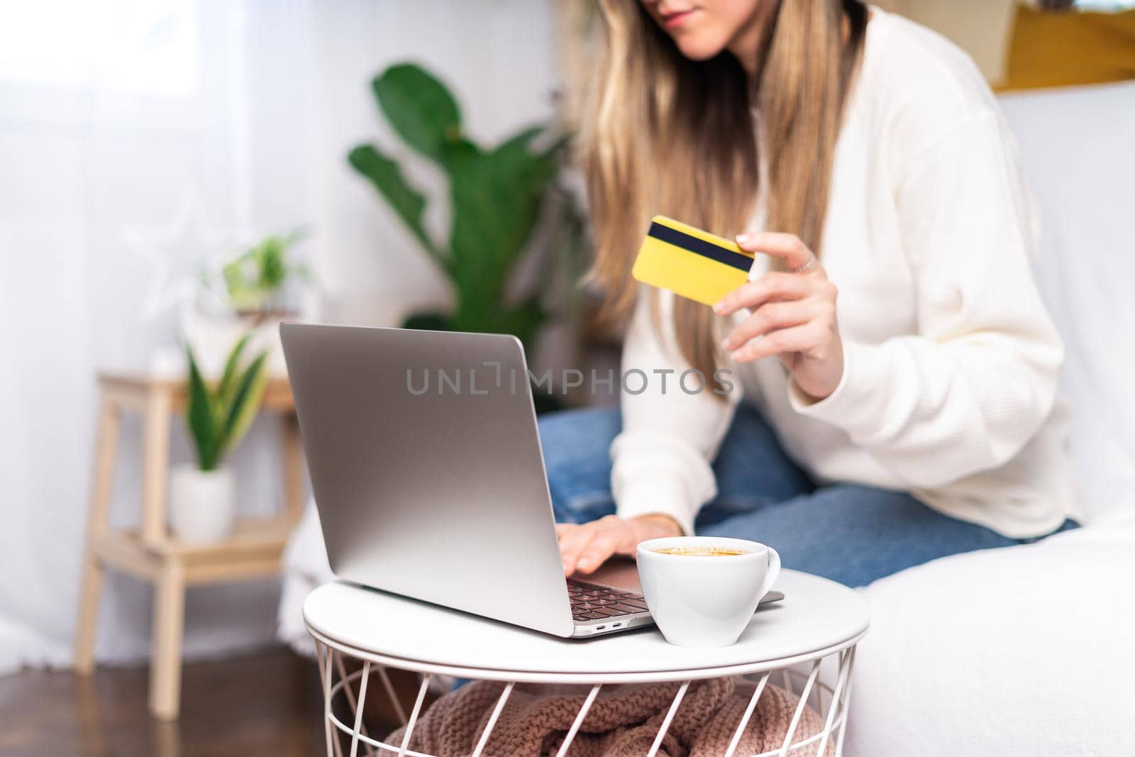 Freelance woman with credit card and coffee cup typing at laptop and online shopping, sitting on the couch. Happy girl woking from home office. Distance learning online education and work.Focus on mug