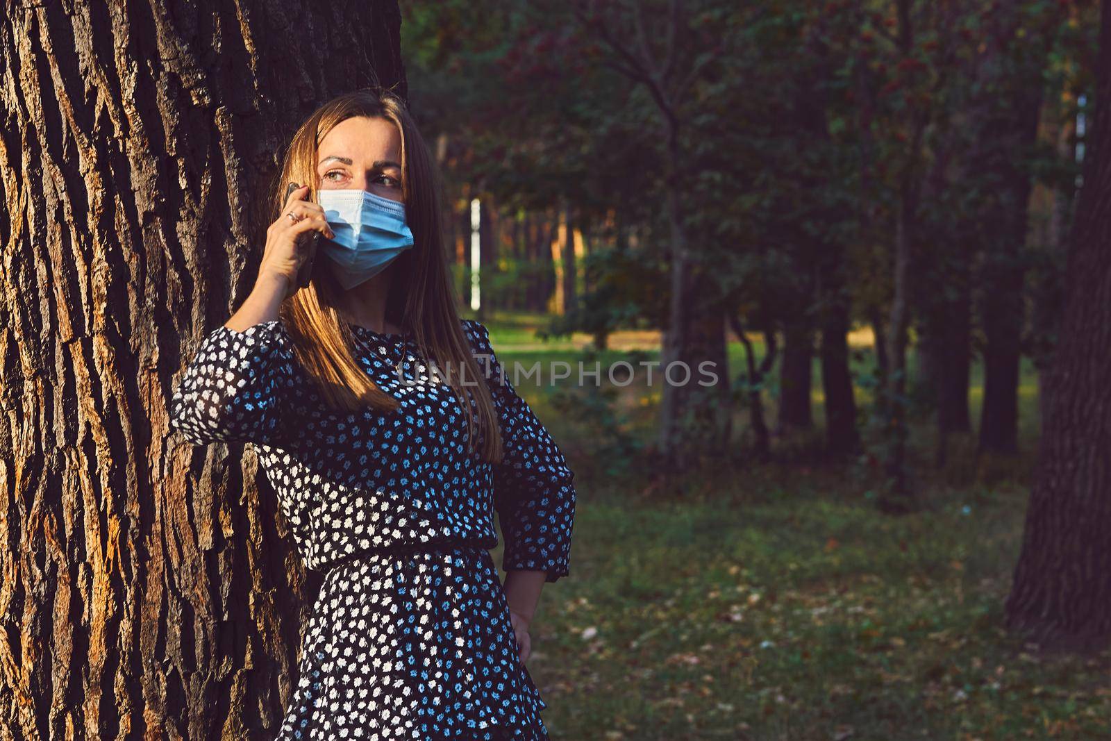 Pretty woman in a mask talking on the phone leaning against a tree in the park by jovani68