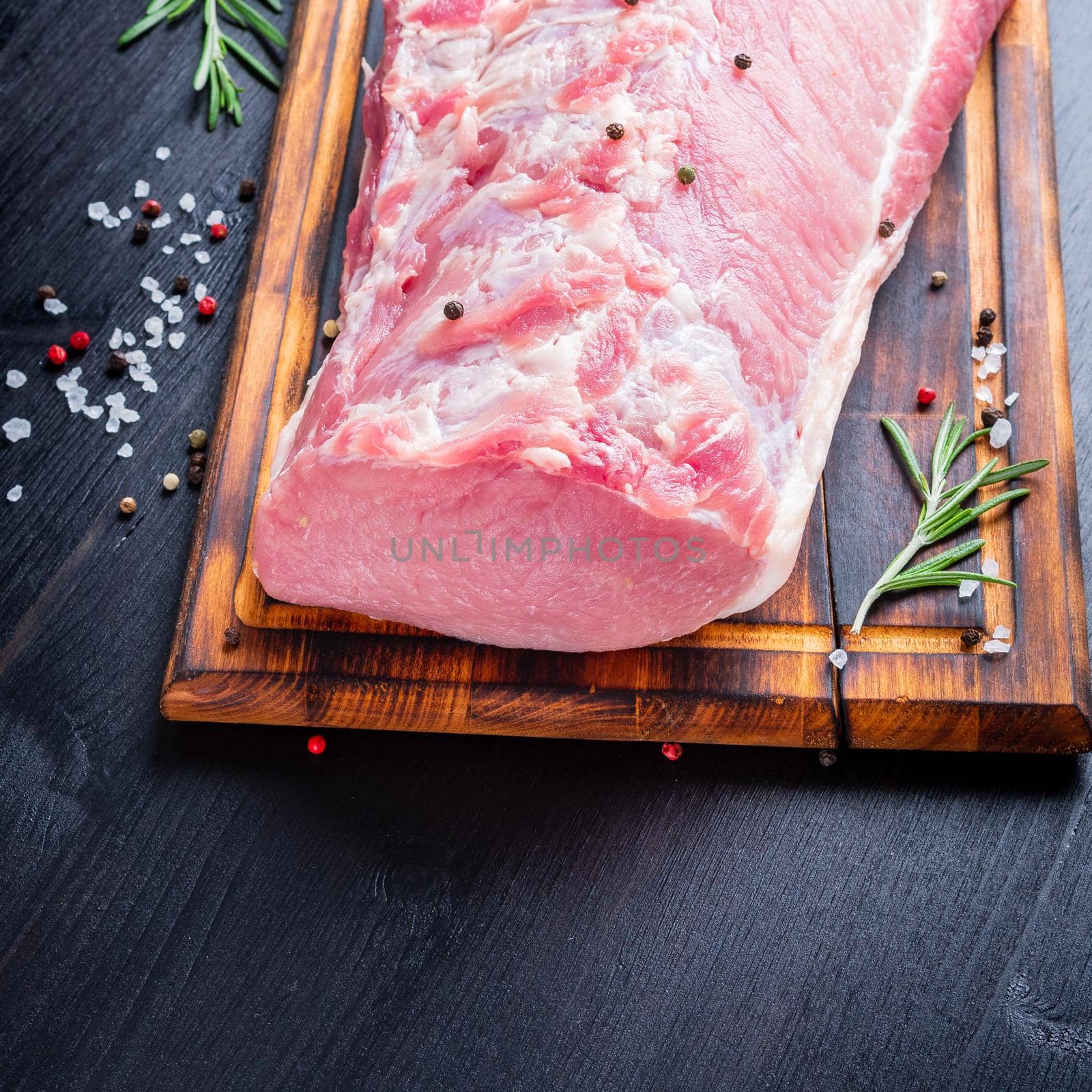 Big whole piece of pork meat with seasoning on chopping board on dark background with rosemary, seasonings, side view.