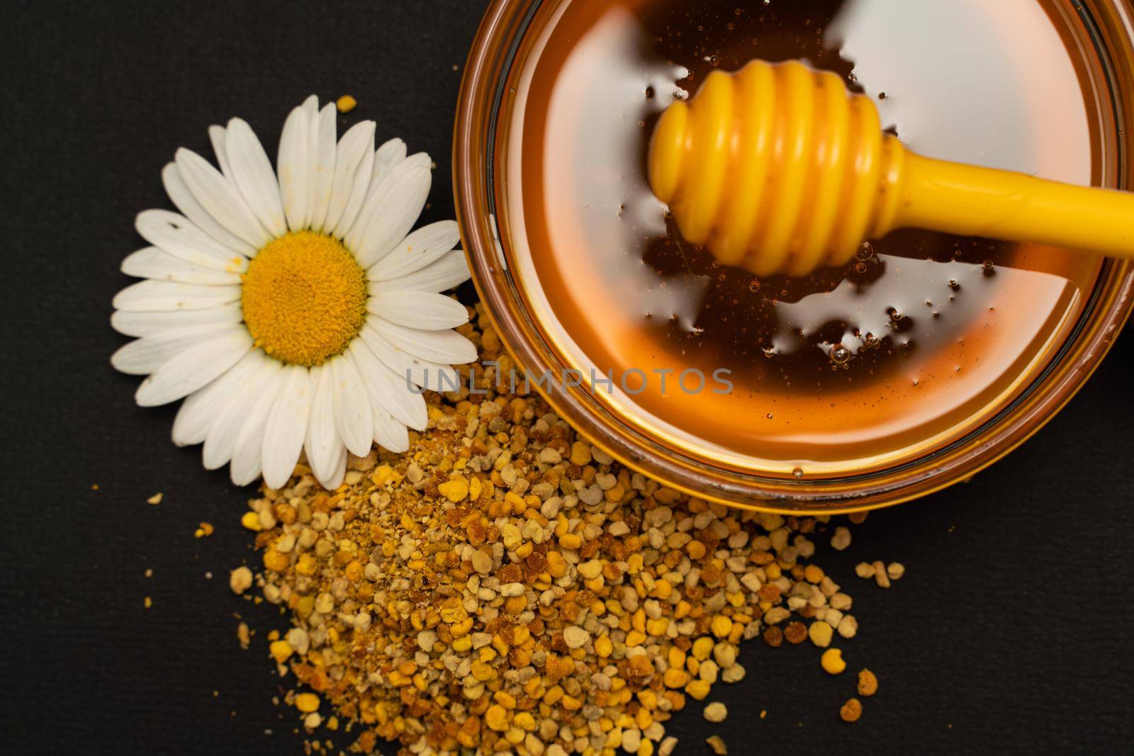 A yellow spoon of honey dipped in a jar of honey and bee bread scattered on a black background decorated with live chamomile. Honey dripping around, nice and inviting photos. Healthy food concept. by Matiunina