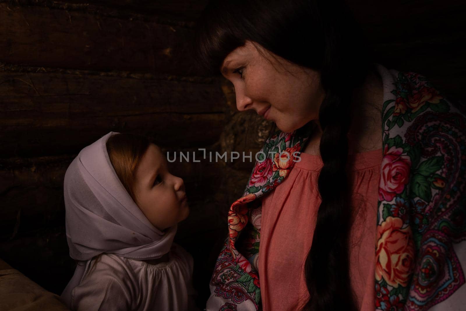a woman and a baby in Russian folk costumes in a bast hut look at each other. Mom smiles at her beloved daughter. The girl looks at her mother enchanted