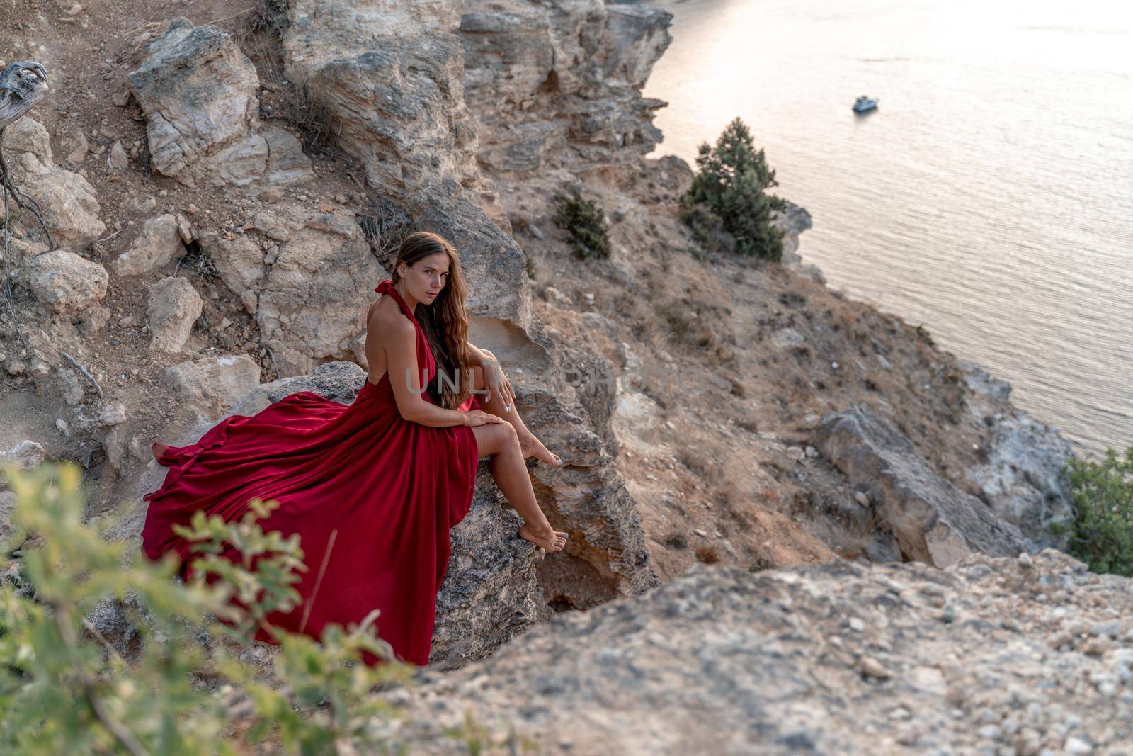 A girl with loose hair in a red dress sits on a rock rock above the sea. In the background, the sea. The concept of travel