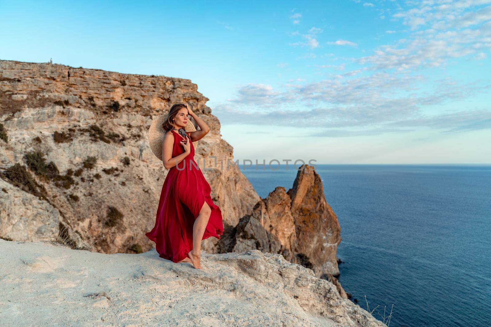 A girl with loose hair in a red dress stands on a rock rock above the sea. In the background, the sea and the rocks. The concept of travel.