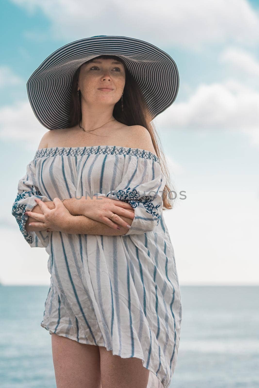 A girl with brown hair and freckles on her face and shoulders in a big blue hat with a large brim and a cape made of cotton fabric on the beach by the sea looks into the distance by olex