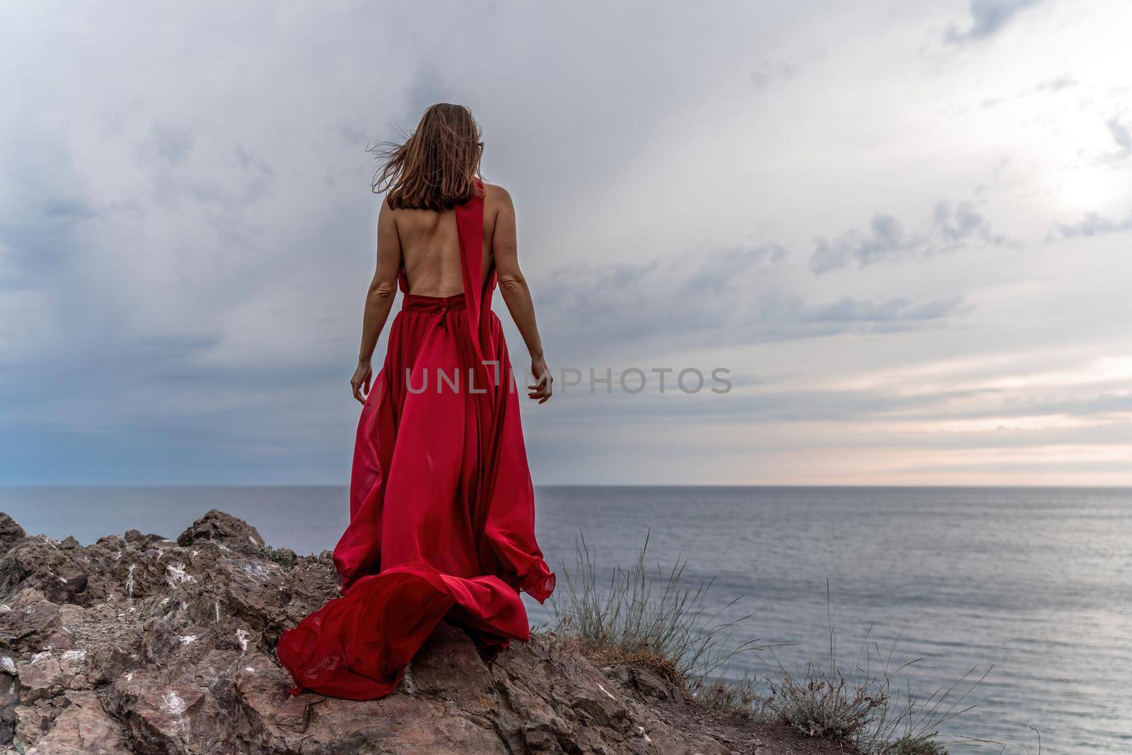 A woman in a red dress stands above a stormy sky, her dress fluttering, the fabric flying in the wind